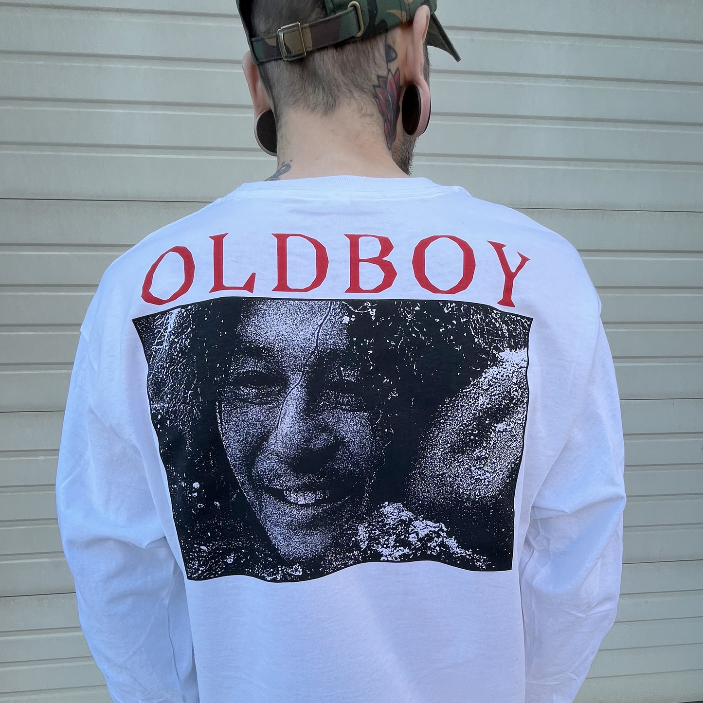A zoomed in photo of the back of the shirt being worn by a model. "Oldboy" is printed in red across the shoulder blades. The text is large and centered over a highly stylized halftone newspaper print photo of the main character.