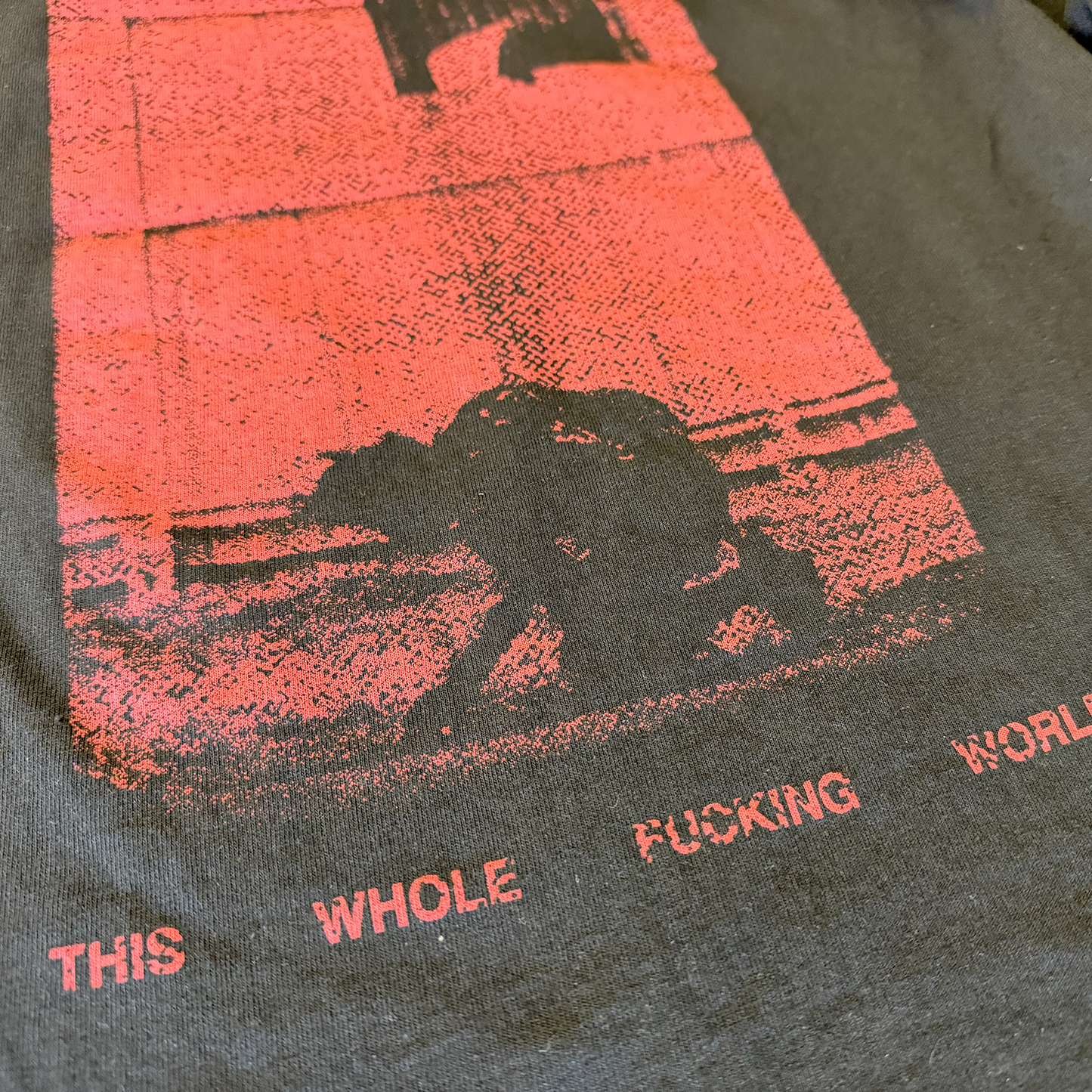 Zoomed in detail of the black version of the shirt. It has a distorted red image of the main character kneeling down to the ground, and it reads "Our love can destroy this whole fucking world"