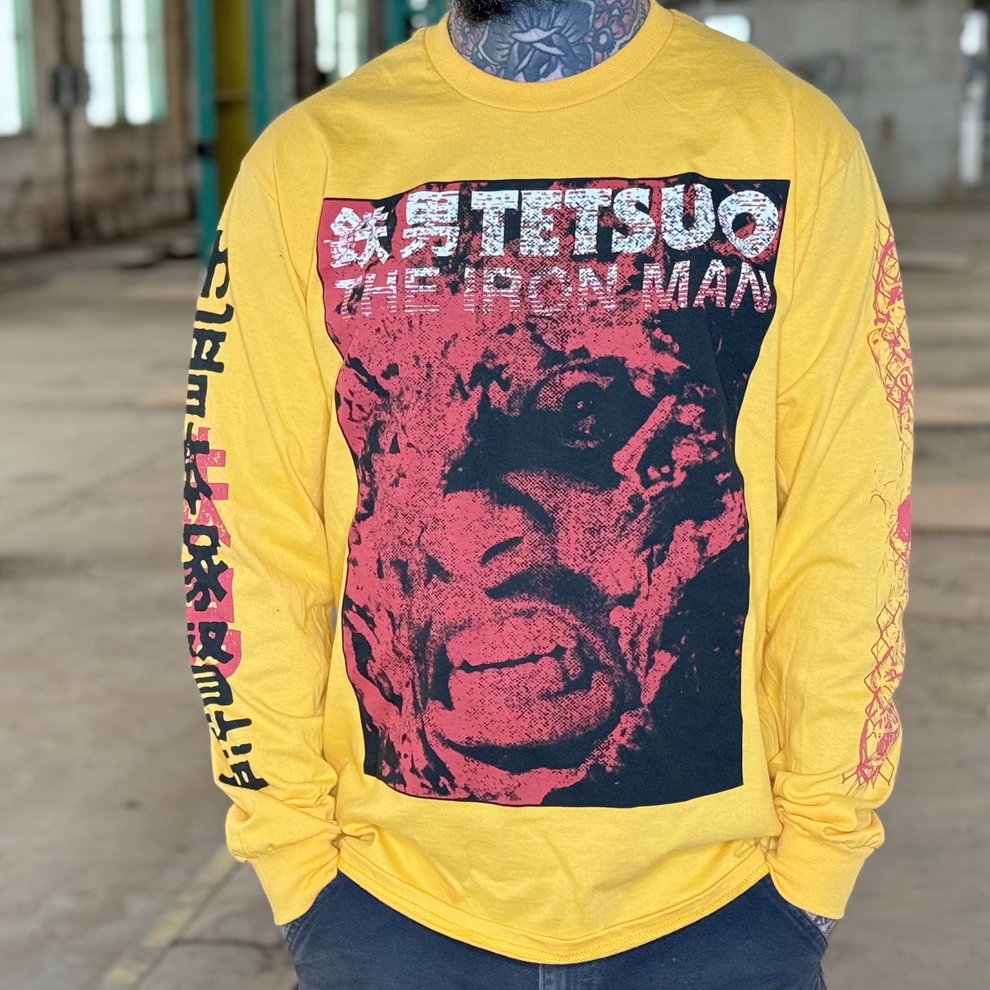 A model wearing the yellow version of the shirt. The picture features the front of the image, with a distorted picture of the main character printed in black and red. The front also reads "Tetsuo The Iron Man" in white.